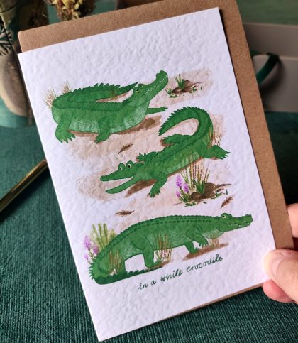 Hand holding a card of 3 cheeky saltwater crocodiles and the hand written note in a while crocodile on the front with Kraft envelope.