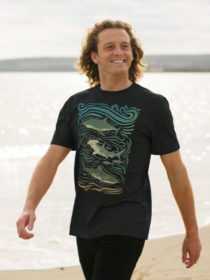 Man walking on sandy beach smiling wearing a black t-shirt with Curious Kin's shark fun facts design printed on it.