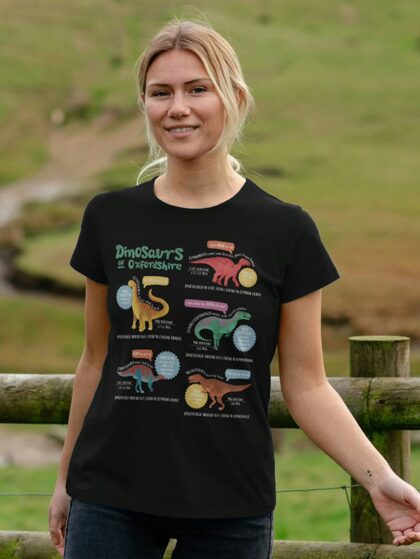 Woman stood in front of a fence wearing a black t-shirt with Curious Kin's dinosaurs of Oxfordshire design