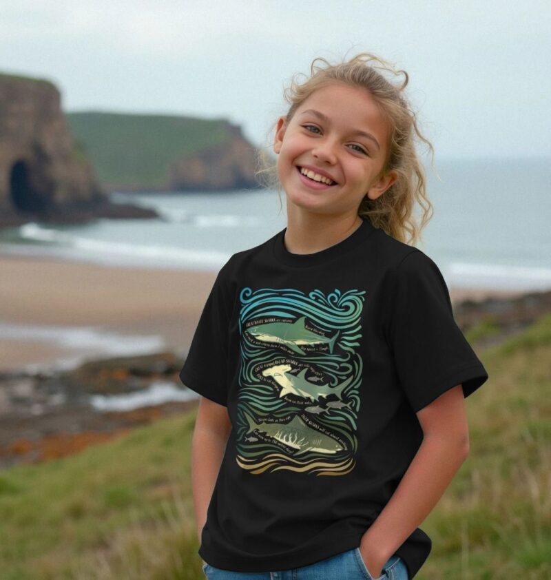 Girl wearing black tshirt with Curious Kin's great sharks fun fact design printed on it hearing great white shark, greater hammerhead shark and tiger shark by the seaside on a grassy hill