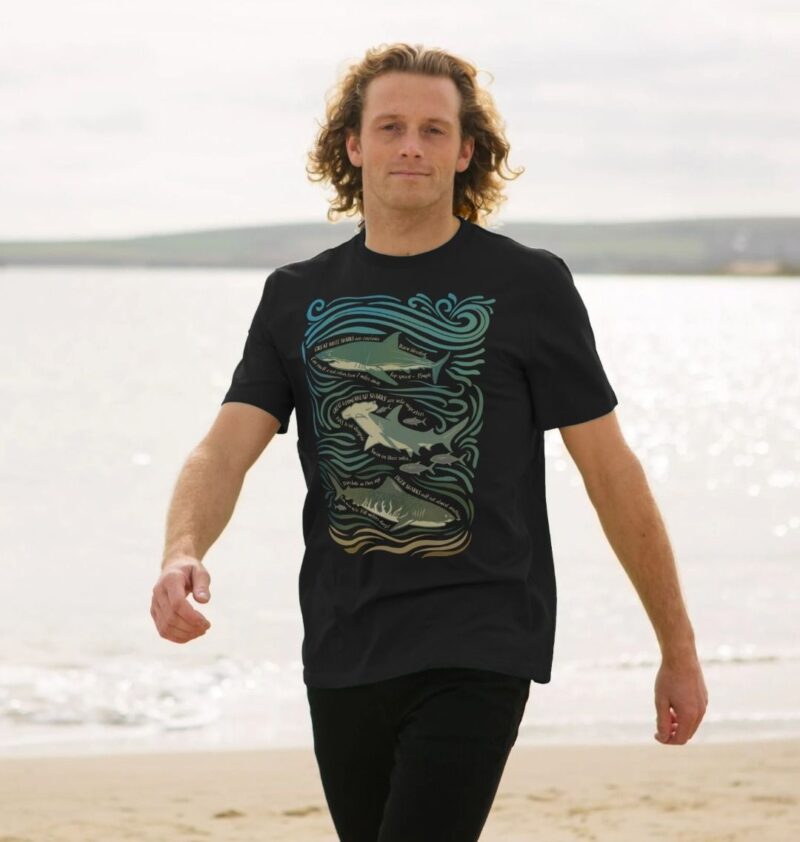 Man walking along a sandy beach wearing black tshirt with Curious Kin's great sharks fun fact design printed on it hearing great white shark, greater hammerhead shark and tiger shark