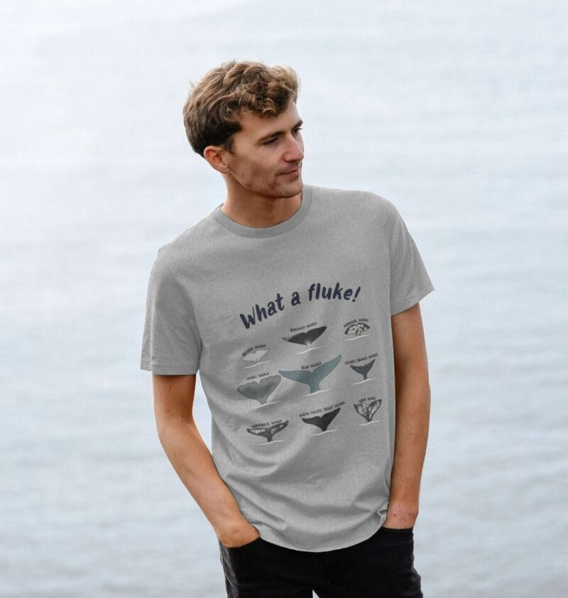 Man with hands in pockets standing by the sea wearing a grey regular fit t-shirt with 9 whale fluke tails and the title what a fluke!