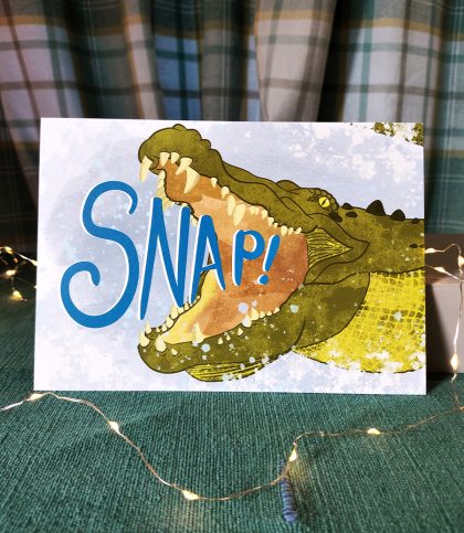 A5 landscape print of an illustration of a saltwater crocodile lunging out of the water and the words Snap! in its mouth