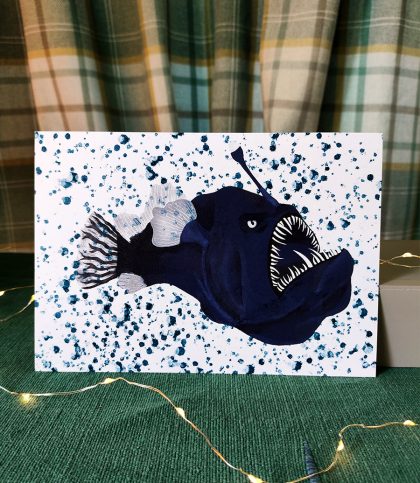 An a5 print of an illustration of a seadevil anglerfish in blue surrounded by paint spatters.