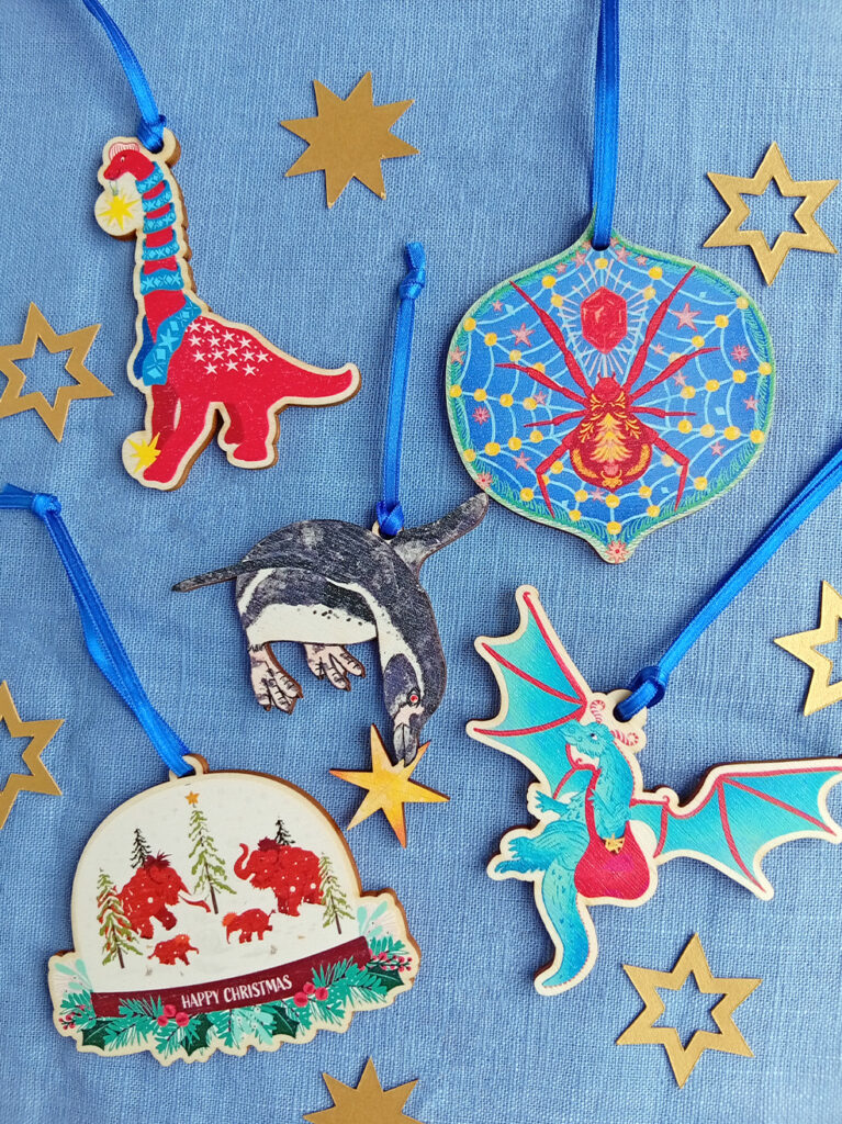 Curious Kin's mixture of illustrated wooden christmas decorations: a christmas brachiosaurus with scarf and star, a christmas spider, a penguin with star, a christmas dragon, mammoth snow globe