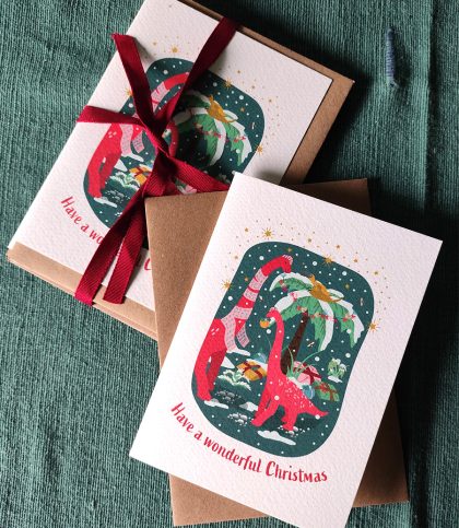 A pair of red brachiosaurus dinosaurs decorate snowy palm trees with a pesky pteradactyl sitting on top and the words Have a wonderful Christmas below. With bundle of cards tied up in red ribbon and matching kraft envelopes.