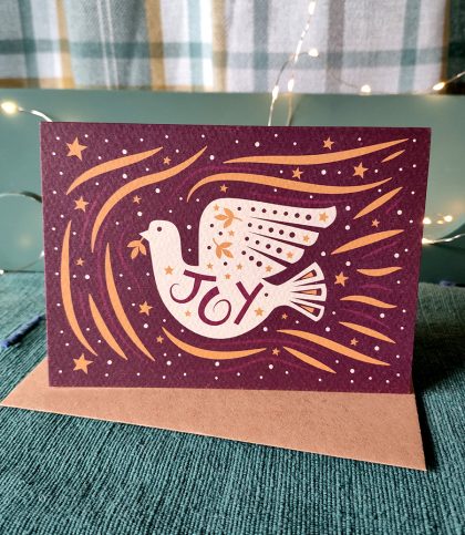 Folk style white dove with hand drawn joy on its tummy and gold stars sitting on a Kraft envelope.