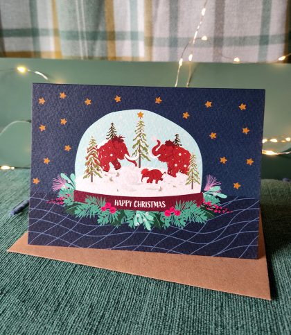 Mammoth family snowglobe with wreath and happy Christmas greeting on a dark blue background. Landscape A6 card sitting on top Kraft envelope.
