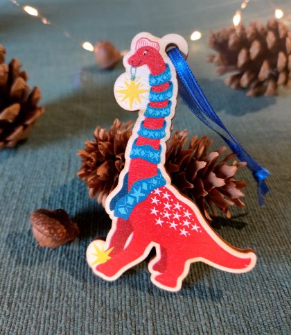 A shaped wooden tree decoration with ribbon tie of a red brachiosaurus holding a bright star in its mouth with a blue scarf wound round its neck and a star at its feet.