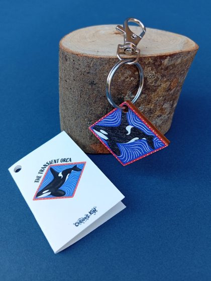Keyring with wooden die-cut diamond charm of a transient orca hanging from a wood block and mini fun fact card lying next to it.