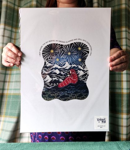 Hands holding up an A3 print of a woman explorer in a sleeping bag on mountain top with starlight and original quote.